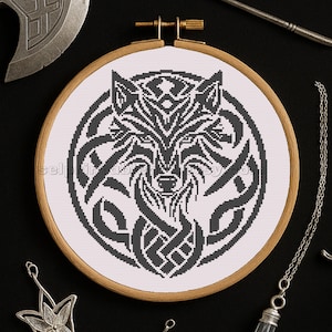 May include: A black and white cross-stitch wolf design in a wooden embroidery hoop. The wolf is surrounded by Celtic knot patterns. The background is white, and the hoop is set against a black surface. The design is detailed and symmetrical.