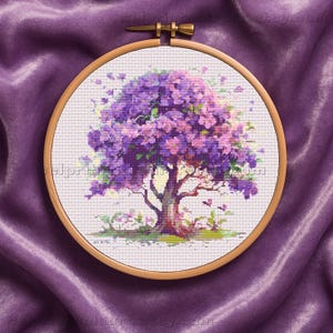 Puede incluir: Bordado de punto de cruz de un árbol con flores moradas y rosas, en un aro de madera. El árbol tiene un tronco marrón y hierba verde en su base. El fondo es una tela de satén morado.
