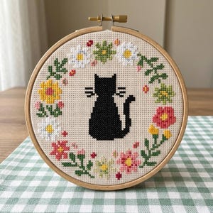 May include: A circular wooden embroidery hoop containing a cross-stitch design on beige fabric. The centre features a solid black cat silhouette surrounded by a colourful wreath of embroidered flowers in shades of white, yellow, pink, and green.