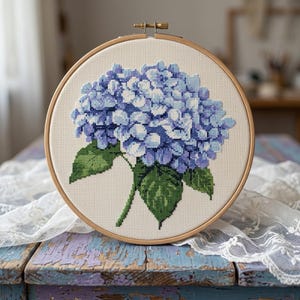 Può includere: Ricamo a punto croce di un fiore di ortensia blu in un telaio di legno. Il fiore presenta diverse sfumature di blu e foglie verdi. Il ricamo è realizzato su un tessuto color crema.