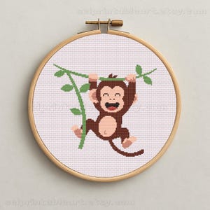 May include: A cross-stitch embroidery hoop featuring a smiling brown monkey hanging from a green vine. The monkey has a light brown face and belly, with a wide, happy grin. The background is white, and the hoop is made of wood. The text "selprintableart.etsy.com" is at the bottom.