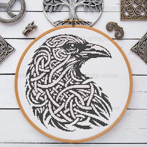 May include: A cross-stitch embroidery of a raven's head, featuring intricate black and white knotwork. The artwork is set in a wooden embroidery hoop. Decorative metal pieces surround the hoop on a white wooden surface.