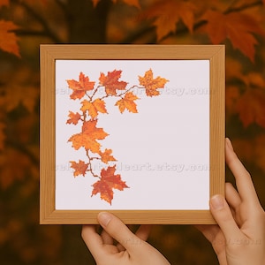 Könnte beinhalten: Ein gerahmtes Kreuzstichbild mit einem Zweig herbstlicher Ahornblätter in Orange- und Gelbtönen. Die Blätter sind auf weißem Hintergrund gestickt und in einem hellbraunen Holzrahmen gefasst. Das Kunstwerk wird von zwei Händen gehalten.