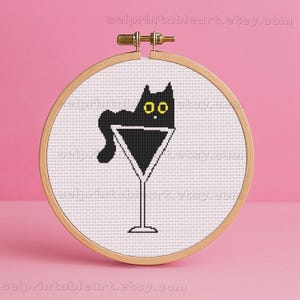 May include: A cross-stitch design shows a black cat in a martini glass, set within a wooden embroidery hoop. The cat has yellow eyes and is on a white background. The text "selprintableart.etsy.com" is visible.