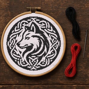 May include: A black and white wolf design cross-stitch in a wooden embroidery hoop. The wolf is surrounded by Celtic knotwork. Black and red embroidery floss and a needle are next to the hoop on a wooden surface.