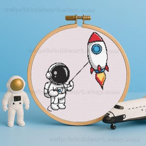 May include: A cross-stitch embroidery hoop featuring a pixelated astronaut holding a rocket on a string. The rocket has red, white, blue, and yellow details. A small astronaut figurine and a toy space shuttle are also in the image.