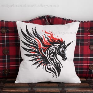 May include: White square pillow with a black, red, and gray cross-stitch design of a winged unicorn. The unicorn has a long horn and a flowing mane. The pillow is on a wooden surface with a red and black plaid background.