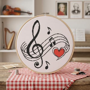May include: A circular wooden embroidery hoop containing a white fabric piece with a cross-stitch design. The design features a large black treble clef, black musical notes, and a red heart with a black outline, all centered on the fabric.