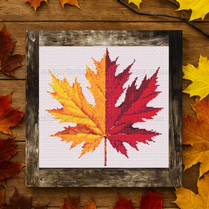 May include: Cross-stitch art of a maple leaf, split vertically with yellow-orange on the left and red on the right. The leaf is framed in a rustic wooden frame, with autumn leaves scattered around.