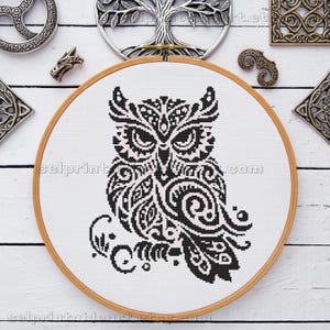 May include: A black and white owl cross-stitch design in a wooden embroidery hoop. The owl features intricate patterns and details. The background is white, and the hoop is surrounded by decorative metal pieces.