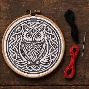 May include: A cross-stitch embroidery project featuring a black and white owl design within a wooden hoop. The owl is surrounded by intricate Celtic knotwork. Black and red embroidery floss and a needle are placed nearby.