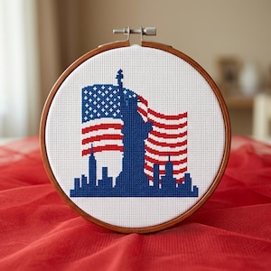 Op de afbeelding: Een borduurring met een patriottisch ontwerp. Het kunstwerk toont het Vrijheidsbeeld en een stadsgezicht in blauw, met de Amerikaanse vlag in rood en wit. De ring is van hout en staat tegen een rode stoffen achtergrond.