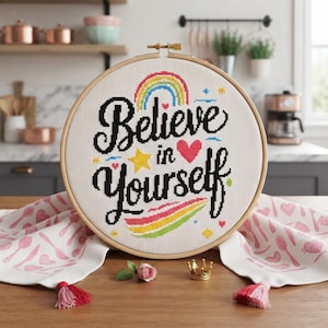 May include: A circular wooden embroidery hoop containing white fabric with a cross-stitched design that reads Believe in Yourself in black cursive lettering, accented by a colorful rainbow, a yellow star, a pink heart, and small geometric shapes.