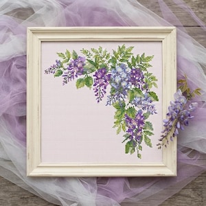 May include: A square cross-stitch embroidery featuring a cluster of purple and blue wisteria flowers with green leaves, set against a light pink fabric background. The piece is enclosed in a distressed off-white wooden frame with a square profile.