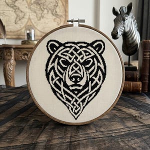 May include: A circular wooden embroidery hoop containing a piece of off-white fabric with a black cross-stitch design of a bear head. The bear is depicted with intricate, interwoven knotwork patterns forming its facial features and ears.