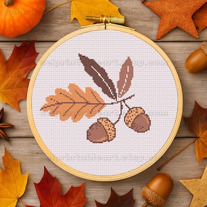 Puede incluir: Un bastidor de bordado de punto de cruz con un diseño de temática otoñal. El diseño incluye un grupo de bellotas y hojas en tonos marrones y naranjas. El bordado está sobre un fondo blanco, rodeado por un aro de madera.
