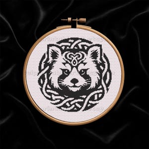 以下が含まれることがあります： ケルトノットデザインのレッサーパンダのクロスステッチ刺繍。パンダは白黒で、木のフープに額装されています。背景は黒いシルクのような生地です。