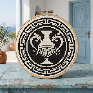 May include: A circular wooden embroidery hoop containing a cross-stitch pattern of a black and white Greek-style amphora vase. The design features a central vase surrounded by a geometric meander border, all rendered in high-contrast black thread.