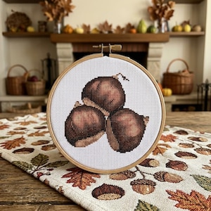 Può includere: Un telaio da ricamo circolare in legno contenente un disegno a punto croce di tre castagne marroni. Le castagne sono realizzate con fili di varie tonalità di marrone su tela Aida bianca, con una vite di tensione in metallo.