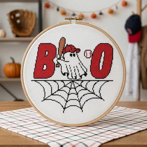 Op de afbeelding: Een ronde houten borduurring met witte stof en een kruissteekontwerp. Het ontwerp toont de rode letters B en O, een wit spookje met een rode honkbalpet dat een bruine knuppel vasthoudt, een honkbal en een zwart spinnenweb.