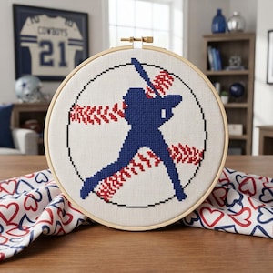 Puede incluir: Un bastidor de bordado circular de madera que contiene un diseño de punto de cruz sobre tela blanca. El diseño presenta la silueta azul oscuro de un jugador de béisbol bateando, superpuesta a un patrón de costuras rojas en un círculo negro.