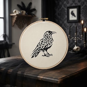 May include: A circular wooden embroidery hoop containing a piece of off-white fabric. Stitched onto the fabric is a black cross-stitch design of a raven featuring intricate Celtic knotwork patterns throughout its body, wings, and tail feathers.