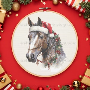 May include: A cross-stitch embroidery of a horse wearing a Santa hat and a festive garland. The horse has a brown and white coat, with a detailed bridle. The embroidery is set in a wooden hoop, surrounded by Christmas decorations.