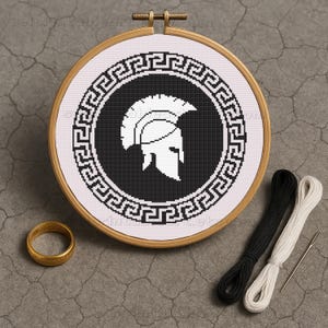 May include: A cross-stitch embroidery in a wooden hoop, depicting a black and white Spartan helmet design within a Greek key border. The image also shows black and white threads, a needle, and a gold ring. The hoop measures approximately 15 cm in diameter.