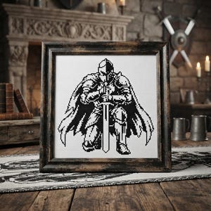 以下が含まれることがあります： 黒と白のピクセルアートで描かれた、剣を立てて跪く鎧姿の騎士のクロスステッチ作品。暗い色調の使い古された木製の正方形のフレームに収められています。騎士は全身鎧を身にまとい、剣を両手で支えるようにして地面に膝をついている様子が表現されています。
