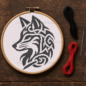 May include: A cross-stitch project featuring a black and white wolf design in a wooden embroidery hoop. The design is detailed with tribal-inspired patterns. Black and red thread, along with a needle, are placed on a wooden surface.