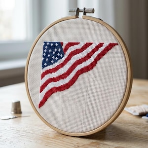 May include: A cross-stitch embroidery of the American flag, featuring red and white stripes and a blue field with white stars. The artwork is displayed in a wooden embroidery hoop, showcasing a craft project.