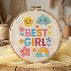 以下が含まれることがあります： カラフルな文字で「BEST GIRL」と書かれたクロスステッチ刺繍枠。デザインには、笑顔の太陽、雲、花、虹が含まれています。枠は木製で、木製の表面に表示されています。