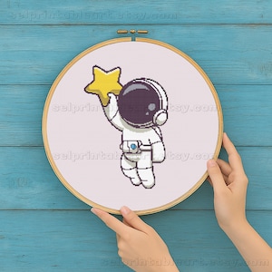 Peut inclure: Un cercle de broderie au point de croix représentant un astronaute pixélisé tenant une étoile jaune. L'astronaute porte une combinaison spatiale blanche et un casque noir. Le texte "selprintableart.etsy.com" est visible.