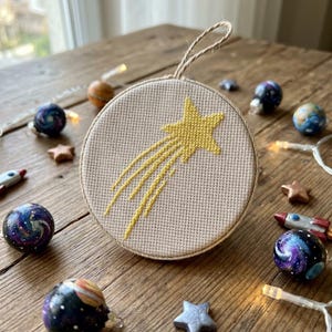 May include: A circular cross-stitch ornament featuring a yellow shooting star embroidered on light beige fabric. The ornament is framed in a wooden hoop and includes a jute twine loop at the top for hanging, showcasing a detailed needlework design.
