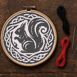 May include: A cross-stitch embroidery hoop with a black and white squirrel design inside a Celtic knot border. The wooden hoop is next to black and red embroidery thread and a needle, on a wooden surface. This craft project is ideal for home decor.