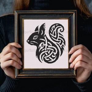May include: Black and white cross-stitch art of a squirrel in a framed square. The squirrel design features Celtic knot patterns. The frame is black with a gold trim. The background is a light pink fabric.