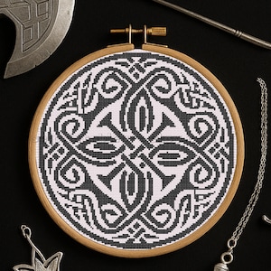 May include: A black and white cross-stitch design in a wooden embroidery hoop. The design features an intricate Celtic knot pattern. The hoop is surrounded by other items, including a silver axe and a necklace.