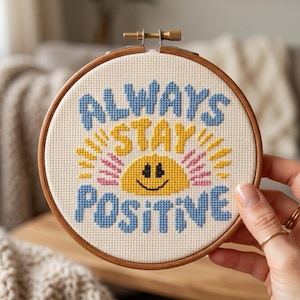 Può includere: Un telaio da ricamo a punto croce con le parole "ALWAYS STAY POSITIVE" in blu e giallo. Il disegno include un sole sorridente con raggi gialli e rosa. Il telaio è in legno e ha una chiusura in ottone.