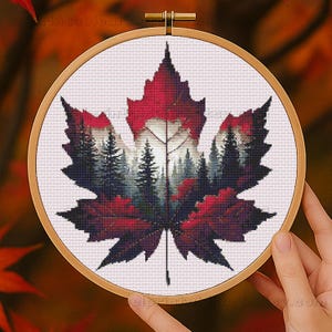 May include: Cross-stitch embroidery of a red maple leaf with a forest scene inside, set in a wooden hoop. The leaf's edges are a deep red, transitioning to lighter shades. The forest features evergreen trees in shades of green and gray.