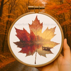May include: A cross-stitch embroidery of a maple leaf in autumn colors, framed in a wooden hoop. The leaf design transitions from red to yellow, with a landscape scene reflected within. The hoop measures approximately 6 inches in diameter.