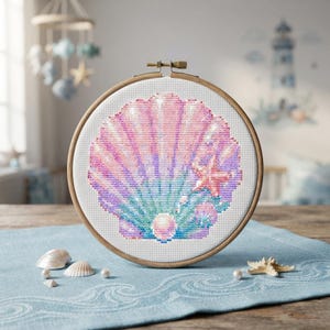 Pode incluir: Um bastidor de bordado circular de madeira contendo um design em ponto cruz de uma concha grande em tons de rosa, roxo e azul. A concha apresenta uma pérola central e uma pequena estrela-do-mar laranja no lado direito sobre tecido branco.