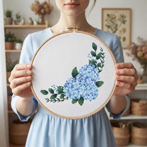 Pode incluir: Um bastidor de bordado circular de madeira contendo tecido branco com um design em ponto cruz de flores de hortênsia azul-claras e folhas verde-escuras. O arranjo floral está posicionado na parte inferior direita da moldura.