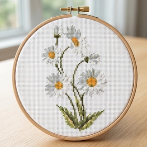 Könnte beinhalten: Kreuzstich-Stickerei mit weißen Gänseblümchen mit gelben Zentren und grünen Stielen und Blättern. Das florale Design befindet sich in einem hölzernen Stickrahmen, ideal für die Heimdekoration oder als handgemachtes Geschenk.