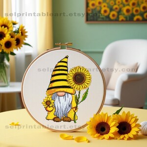 Può includere: Schema di punto croce di un gnomo con un cappello a strisce gialle e nere che tiene un girasole. Lo gnomo è in piedi su uno sfondo bianco.