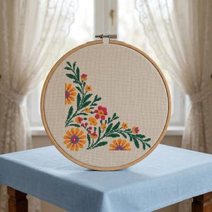 May include: A cross-stitch embroidery in a wooden hoop. The design features a corner arrangement of colourful flowers in shades of orange, pink, and yellow, with green leaves. The fabric is a light beige colour, and the hoop is made of wood.