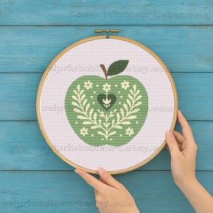以下が含まれることがあります： 緑色のリンゴのデザインが施されたクロスステッチ刺繍枠。ハートと花のアクセントが特徴です。リンゴは白い布の中央に配置され、木製の枠で囲まれています。背景は水色です。