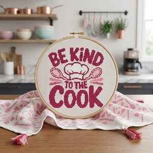May include: A circular wooden embroidery hoop containing white fabric with cross-stitched text that reads BE KIND TO THE COOK, featuring a chef hat and two wooden spoons in dark pink thread, resting on a white cloth with a pink kitchen utensil pattern.
