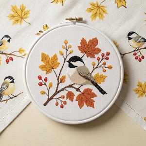 May include: A circular embroidery hoop containing a cross-stitch design of a black-capped chickadee perched on a branch with orange and yellow autumn leaves and red berries, set against a white fabric background with a matching bird and leaf print.