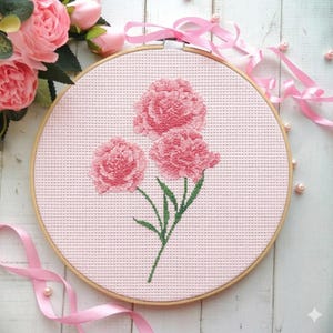 May include: A cross-stitch embroidery hoop featuring three pink peony flowers with green stems and leaves. The flowers are centered on a light pink fabric, set within a wooden hoop. Pink ribbon and faux pearls add a decorative touch.
