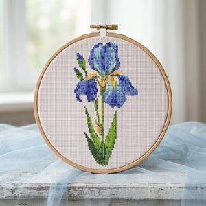 May include: A completed cross-stitch embroidery of a blue iris flower in a wooden hoop. The flower has blue petals with yellow accents and green leaves. The embroidery is set against a white background.